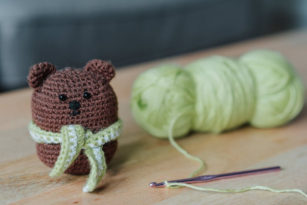 Closeup of small dark brown crochet toy bear and crochet next to light green threads on wooden table in bright room on blurred background
